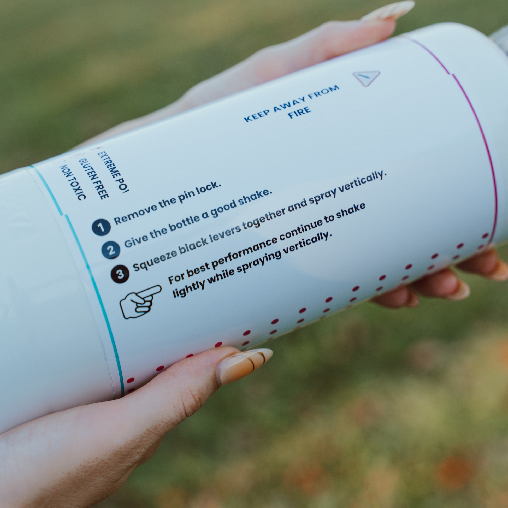 Gender Reveal Double MEGA Powder Blaster instructions shown on white canister displaying step-by-step usage guide: remove pin lock, shake bottle, squeeze levers, and spray vertically for best performance. Close-up view against blurred green background.