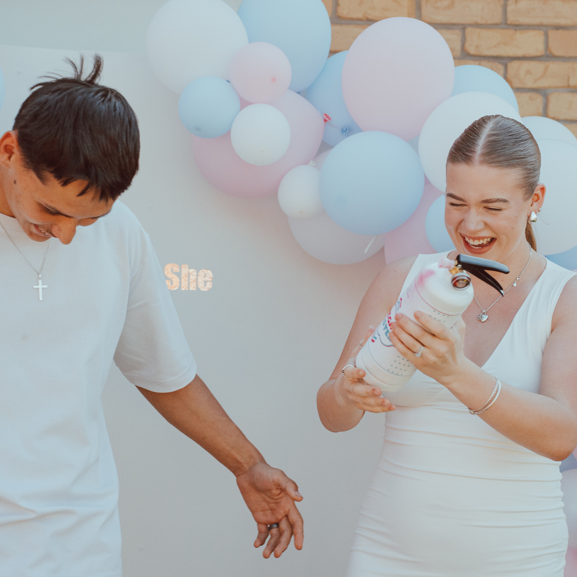 A couple celebrates with a Gender Reveal Double MEGA Powder Blaster against a backdrop of pink and blue balloon arch. The extinguisher is held while they share a joyful moment, illustrating the product's use at an outdoor gender reveal party.
