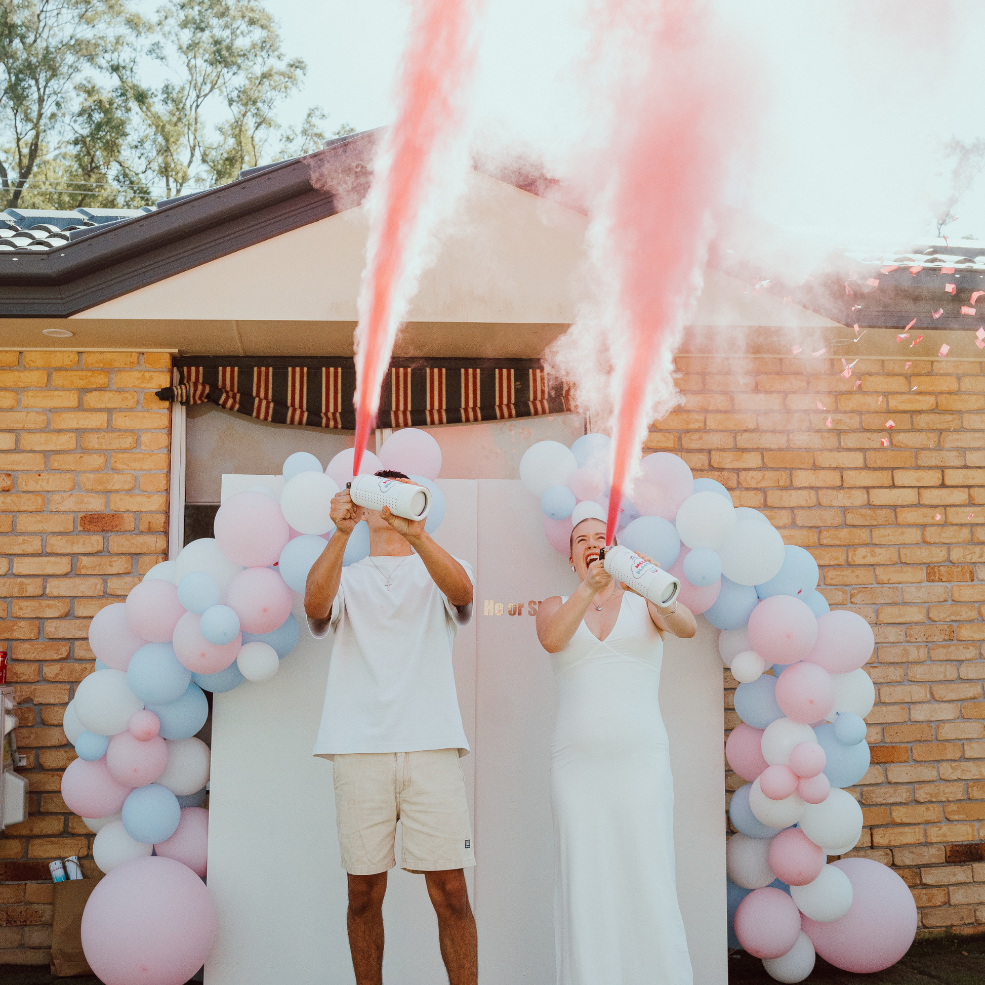 Gender Reveal Double MEGA Powder Blaster creates spectacular pink smoke streams under a decorative balloon arch featuring pink and blue balloons against a brick house backdrop. Two people in white outfits simultaneously activate the powder blasters for a dramatic reveal effect.