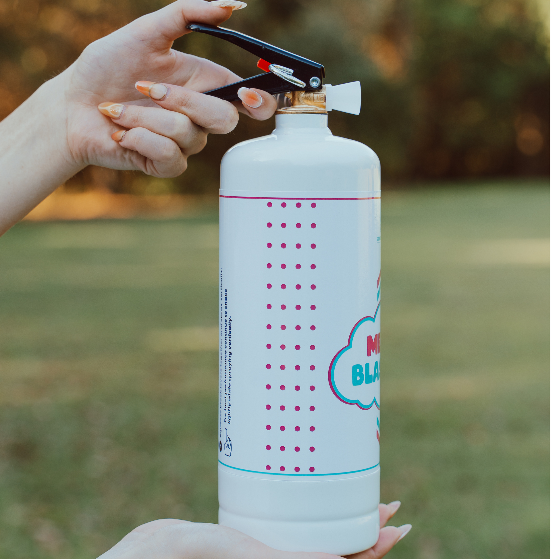 Gender Reveal Double MEGA Powder Blaster/Extinguisher shown as white canister with pink dotted pattern and blue cloud design, held by hands against green outdoor background, demonstrating grip and activation mechanism for gender reveal celebrations