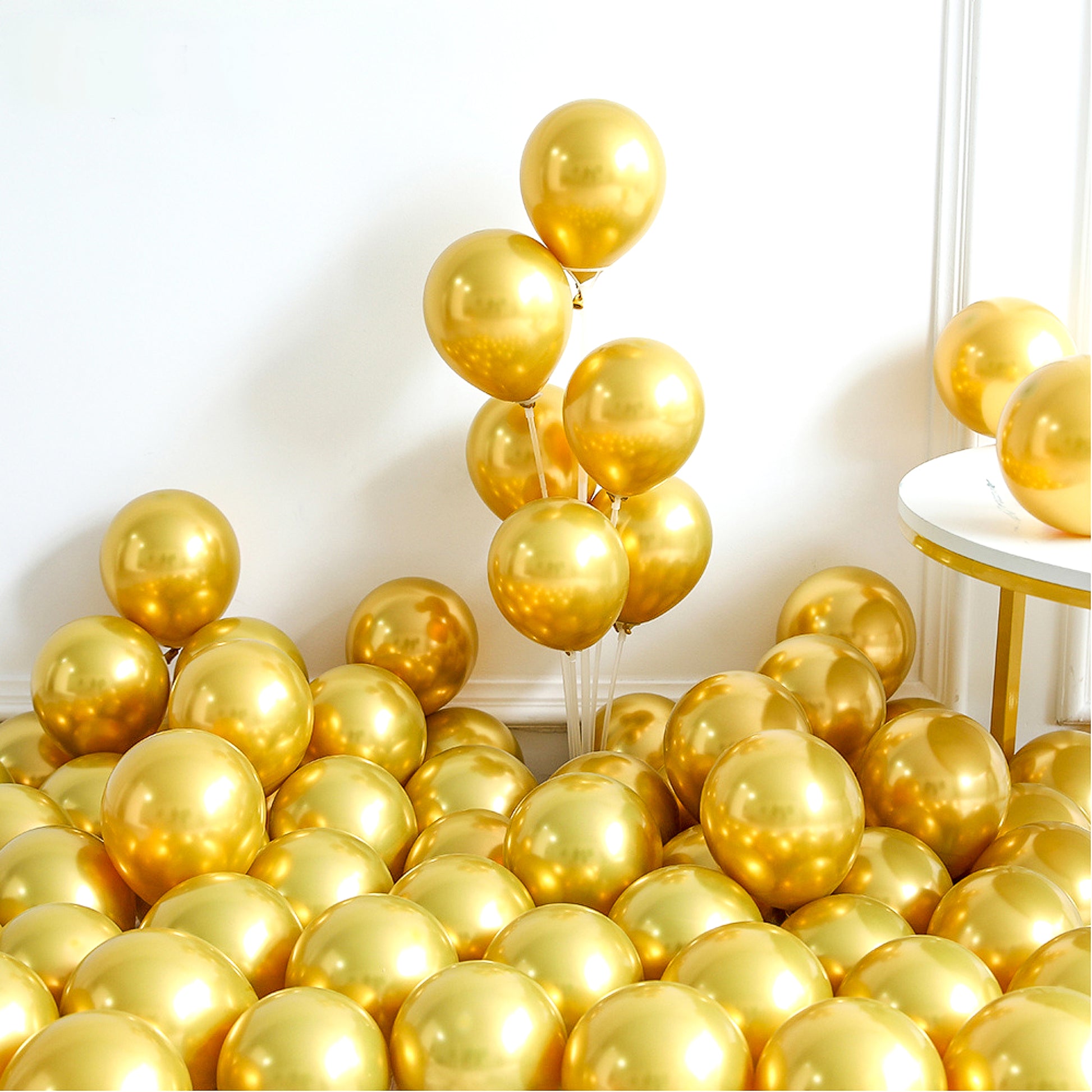 Luxurious display of metallic gold balloons from 148 Piece Universal Gender Reveal/Baby Shower Luxury Balloon Kit arranged artistically against white background, showcasing chrome finish and elegant party decoration setup with round side table