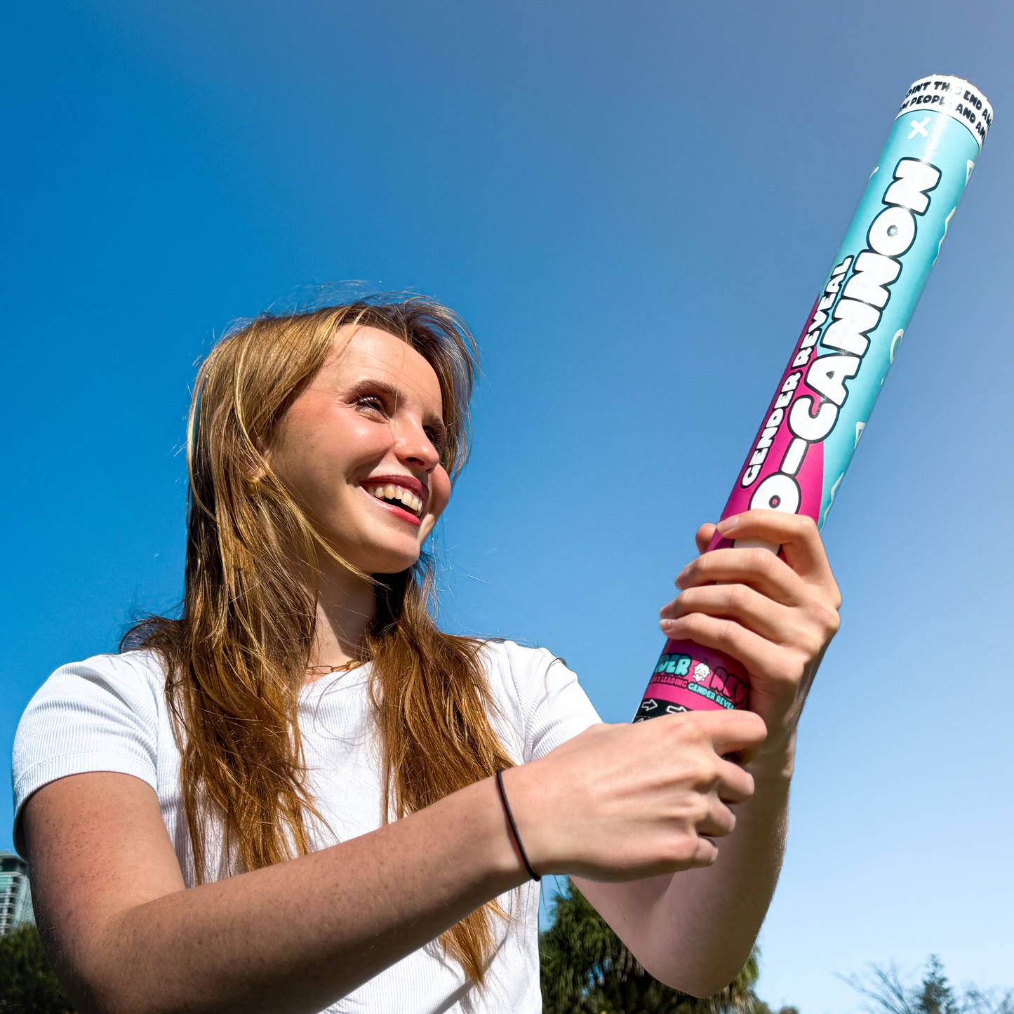Person holding a Gender Reveal Confetti Cannon XL 50cm against bright blue sky, wearing white t-shirt, demonstrating the large size and festive design of the pink and blue cannon outdoors in natural lighting.