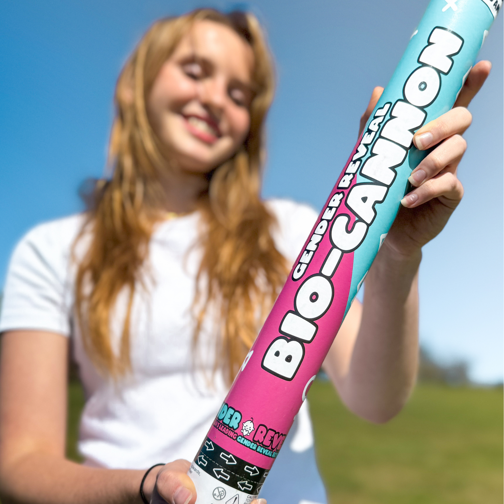 A person in a white t-shirt holding a large biodegradable gender reveal cannon in pink and blue colors against a bright blue sky. Part of the 4 Pack XL Gender Reveal Confetti & Powder Cannon Bundle for celebrating special moments outdoors.