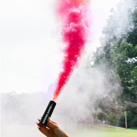 Extreme Gender Reveal Smoke Bomb emitting vibrant pink smoke against a natural outdoor backdrop, demonstrating the product's powerful colored smoke effect during use. Hand holding black smoke emitter canister with dramatic upward smoke plume.