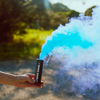 Extreme Gender Reveal Smoke Bomb emitting vibrant blue and white smoke clouds in an outdoor setting, held by a hand against a natural green background, demonstrating the product's dramatic smoke effect for gender reveal celebrations.