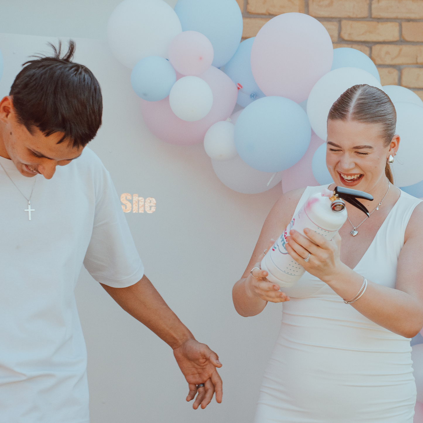 A couple celebrates with Gender Reveal Extinguisher MEGA Powder Blaster against pink and blue balloon arch backdrop. They share a joyful moment holding the white extinguisher canister, with 'She' illuminated text visible in background.