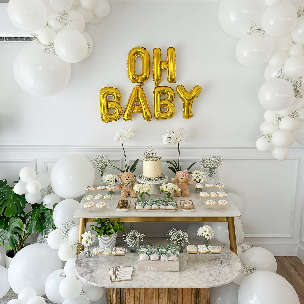 Baby Shower Party Box Set display featuring elegant dessert table with gold OH BABY balloon letters, white balloon arch, teddy bears, and tiered marble tables adorned with treats and floral arrangements, showcasing neutral-toned party decor against a white wall.