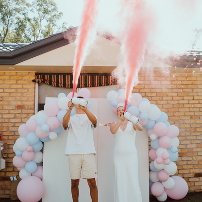 Gender Reveal Extinguisher MEGA Powder Blaster creating dramatic pink powder streams under a decorative balloon arch in pink and blue colors against brick house backdrop, demonstrating product performance during outdoor gender reveal celebration.