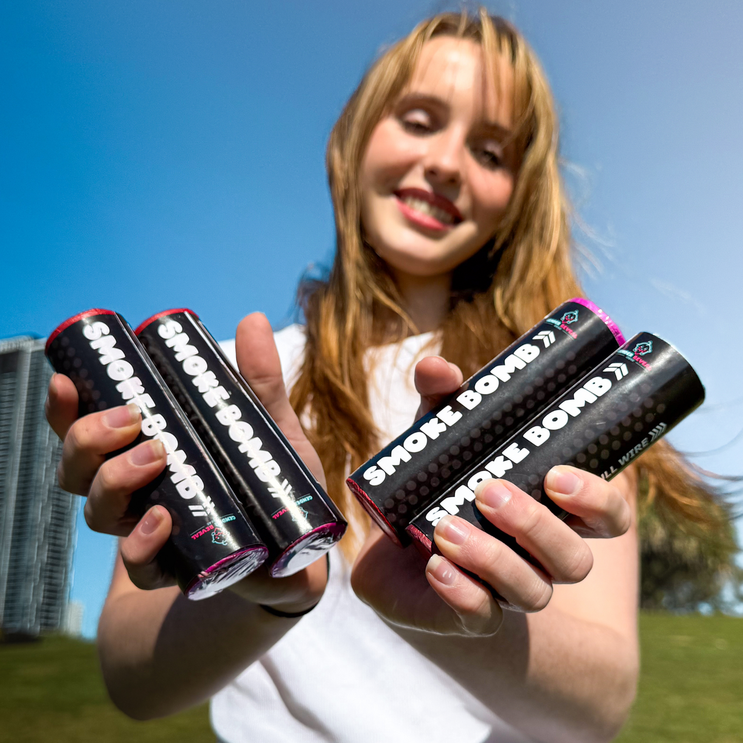 A sunny outdoor product shot of Extreme Gender Reveal Smoke Bombs displayed against a blue sky background, with four black cylindrical smoke emitters featuring white text branding shown in hands wearing white against green grass and modern buildings.