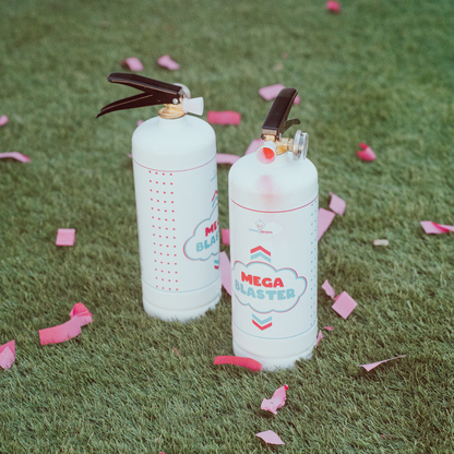 Gender Reveal Extinguisher MEGA Powder Blaster set featuring two white powder cannons with cloud-shaped logos displayed on grass, surrounded by scattered pink confetti petals in a dreamy, soft-focused outdoor setting