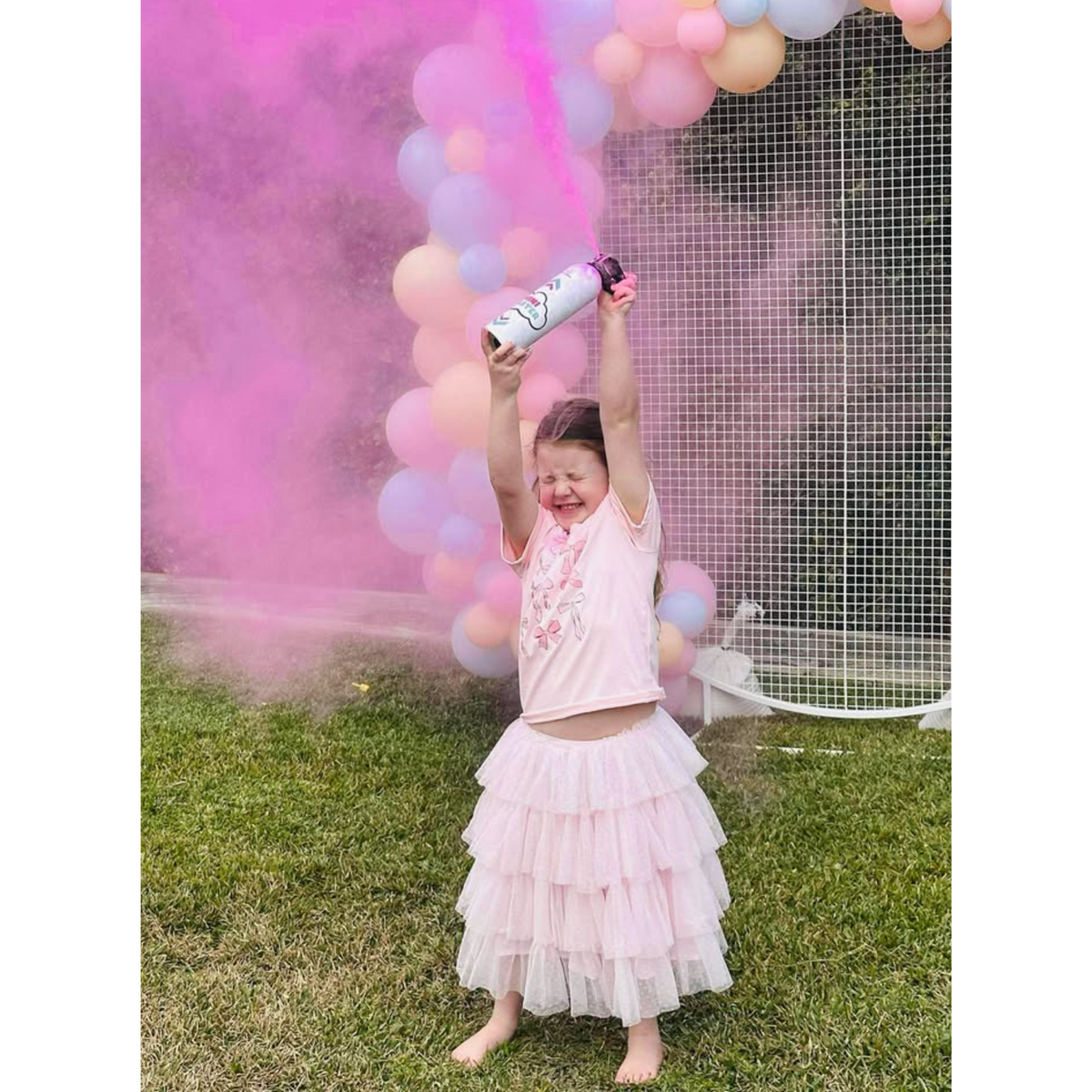 Young girl releasing pink powder from a gender reveal mini extinguisher in front of balloon decorations outdoors