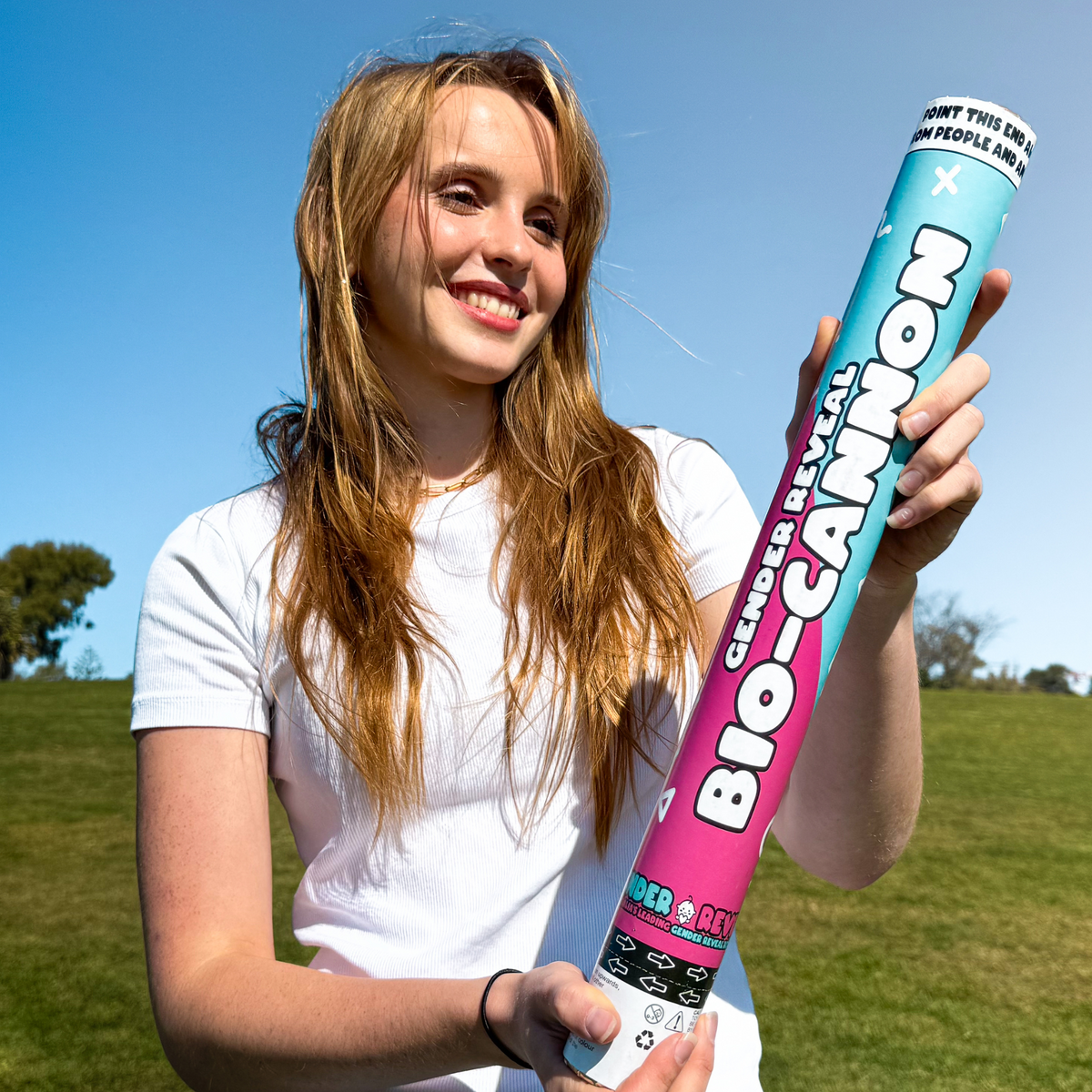 Person in white t-shirt holding Gender Reveal Confetti & Powder Cannon XL 50cm outdoors against blue sky and grassy hill background, showcasing the large blue and pink gradient product tube with excited expression.