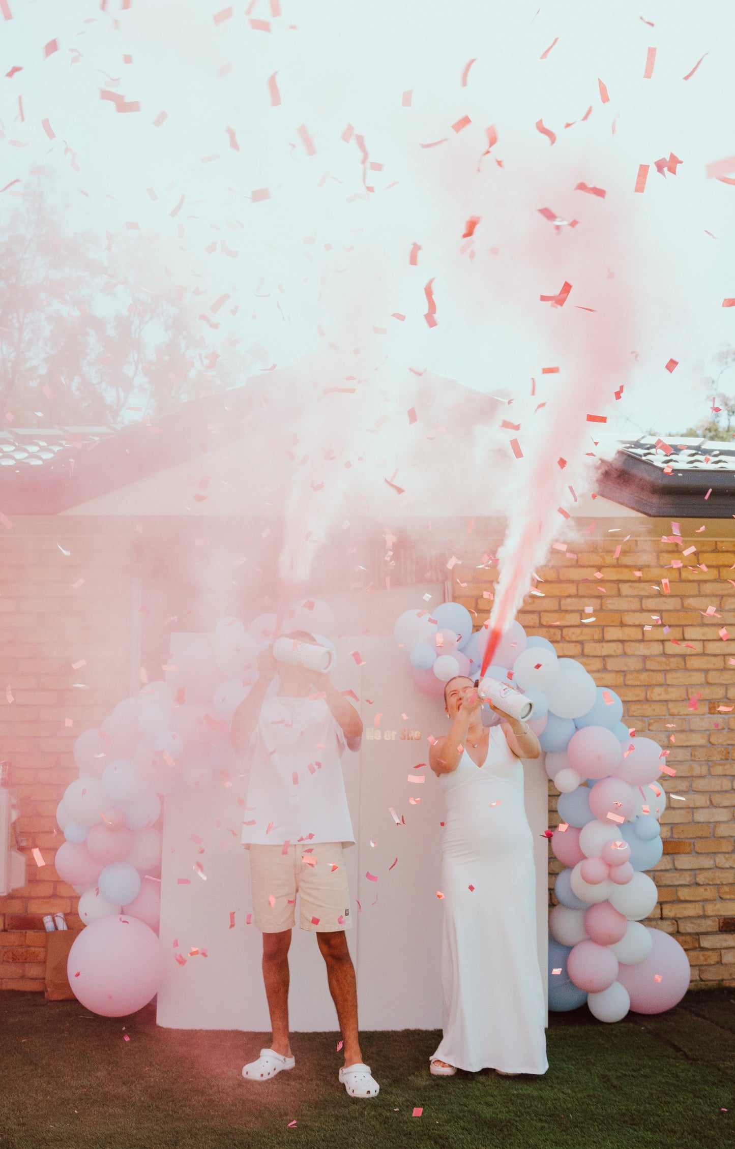 Double MEGA Powder Extinguisher & Gender Reveal Cannon Bundle in action, showing pink powder explosion against brick wall backdrop with decorative pink and blue balloon arch. Powder creates dramatic plume effect while confetti scatters in the air, capturing an exciting gender reveal moment.