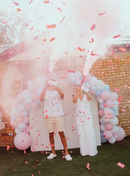 Double MEGA Powder Extinguisher & Gender Reveal Cannon Bundle creating spectacular pink smoke plumes and confetti burst under decorative pink and blue balloon arch. White smoke effect and pink confetti fill the air during outdoor reveal celebration.