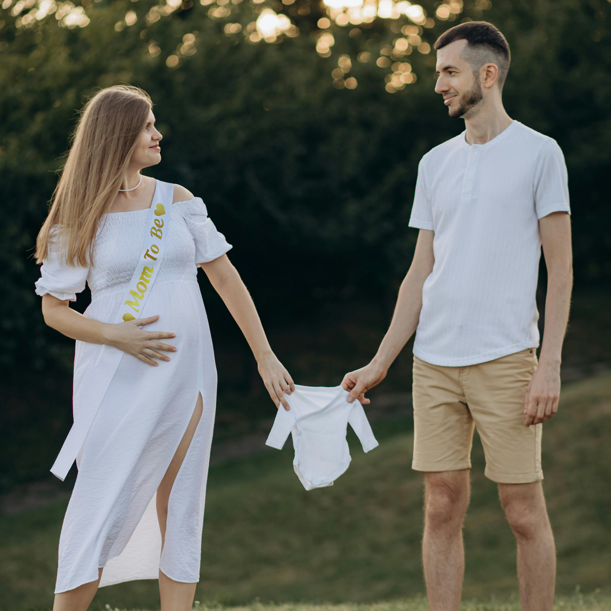 Expectant couple in an outdoor setting at sunset, with the mother wearing a white Mom To Be Gender Reveal Sash and dress, holding her belly, while partner holds a small white baby onesie. Both are dressed in light colors against a natural background.