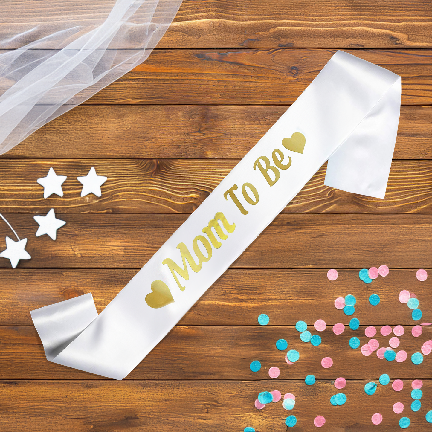 Mom To Be Gender Reveal Sash displayed on wooden background with gold lettering, surrounded by white stars, tulle fabric, and pink and blue confetti scattered decoratively across the surface.