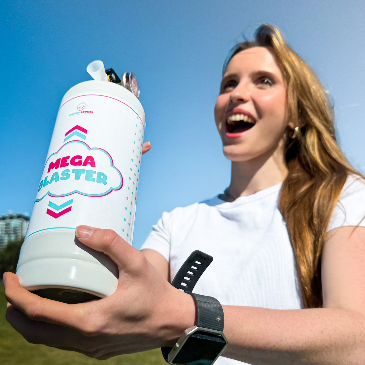 White Quad Gender Reveal MEGA Powder Blaster shown outdoors against blue sky, held by someone wearing white t-shirt and smartwatch. Product features pink and blue chevron design with MEGA BLASTER text on cylindrical white container.